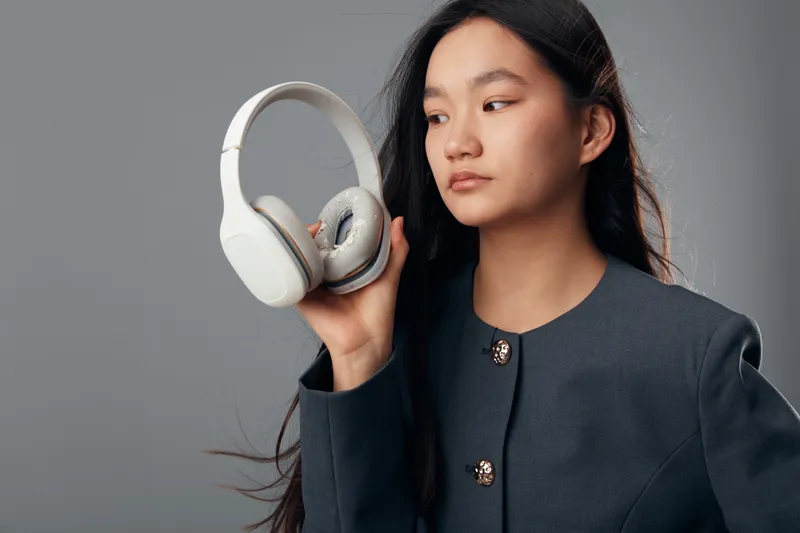 Woman holding wireless headphones close to ear with focused expression standing against gray background. Asian female model with long black hair wearing dark jacket in studio light.