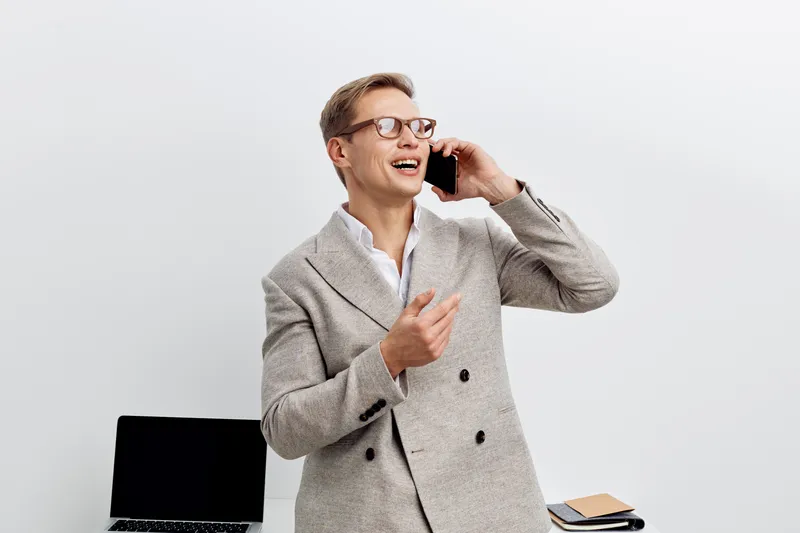 Confident mature man wearing glasses and stylish gray blazer talking on smartphone, smiling, standing in modern office environment with laptop and notepad, professional business look, communication co