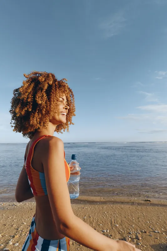 Woman with curly hair on sandy beach, holding a small cup of water and looking toward the ocean. Summer vacation, relaxation, nature and bright sky.