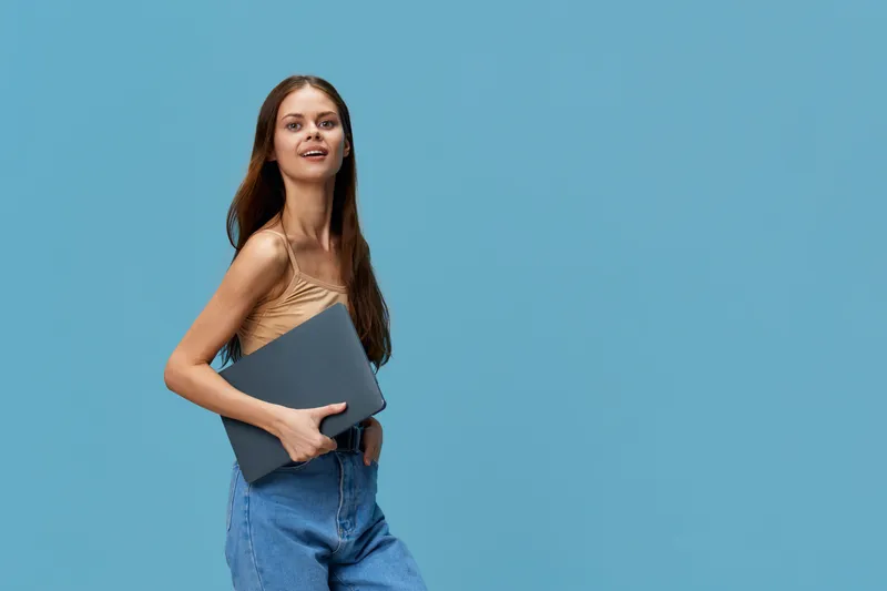 young woman holding laptop wearing casual beige top and blue jeans stands confidently against blue background with a slight smile, embodying modern tech lifestyle