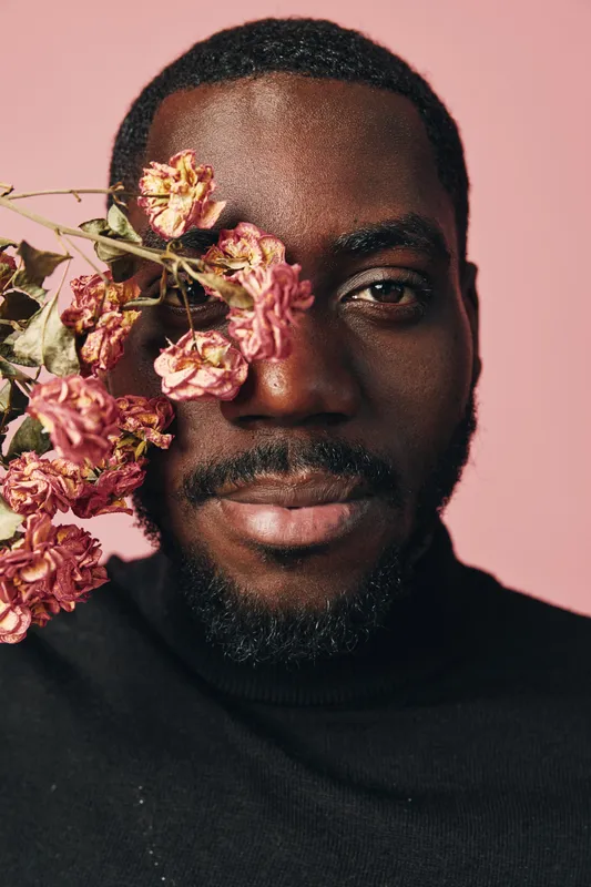 African man with dark skin and beard posing with dried pink flowers covering one eye in creative conceptual visual style on pink background. Portrait with artistic expression and fashion.