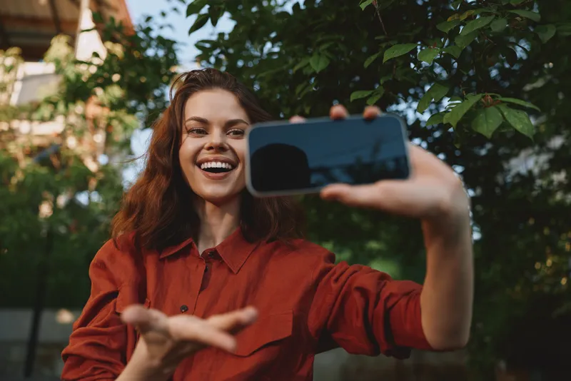 Smiling woman taking a selfie outdoors, showcasing lifestyle and casual fashion. Portrait shot with smartphone, natural light, verdant background, highlighting happiness and confident expression