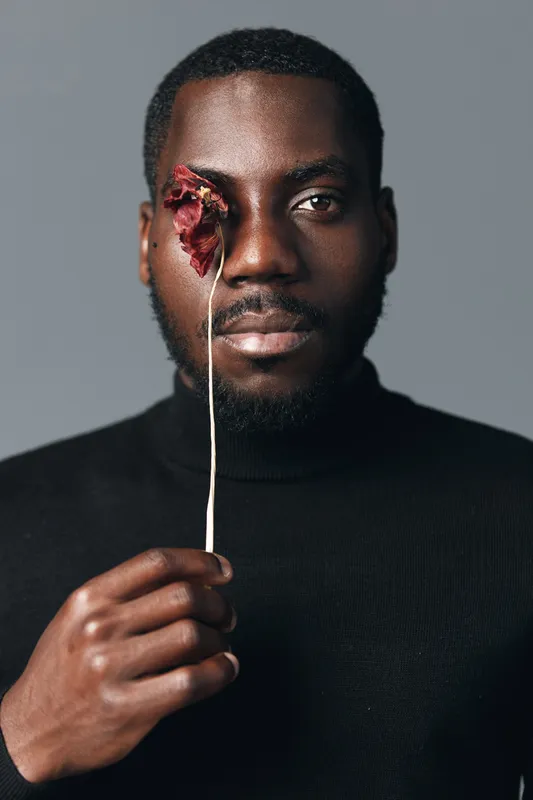 African man with dark skin holding dried flower in front of one eye in creative conceptual visual style. Portrait with black turtleneck and neutral background for artistic expression.