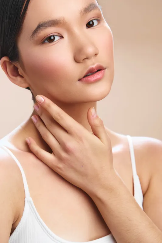 Asian woman with smooth skin gazing softly, her hand near her neck, conveying calm beauty and gentle self care in a clean studio portrait.