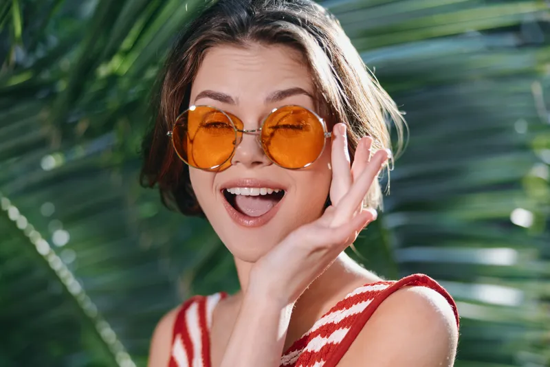 A cheerful woman wearing round orange sunglasses smiles brightly while posing in a sunny tropical setting, capturing a vibrant, carefree vibe and a moment of playful confidence.