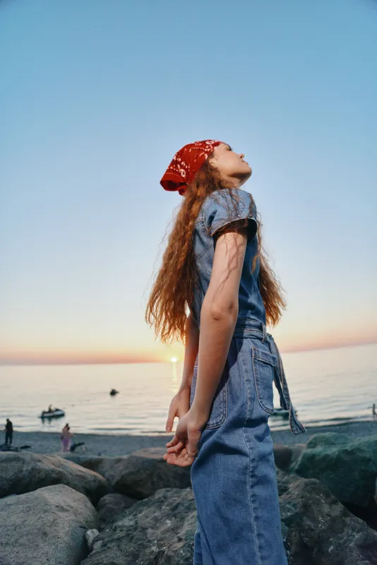 Lifestyle woman in modern boho-western denim street style stands by the seaside during sunset with a red bandana, natural light, and film color atmosphere.