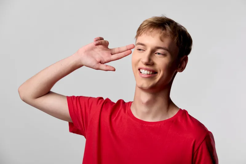 Smiling man in a red shirt makes a light hand gesture beside his head, conveying playful confidence and casual style for lifestyle, fashion and advertising
