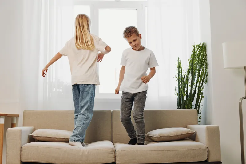 Two playful children jumping on beige sofa in bright living room with plants and natural light. Concept of fun, childhood, and home activity indoors.