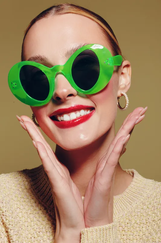 Smiling woman with bright red lipstick wearing large green sunglasses and beige sweater poses with hands near face on neutral background. Concept of fashion, style, and joyful expression.