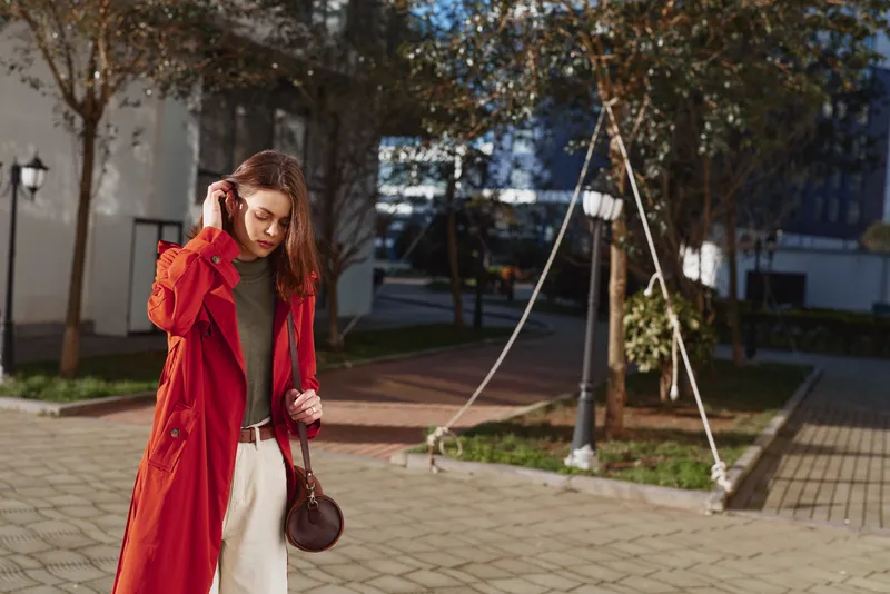 Young woman with long brown hair wears a red trench coat and beige pants while walking outdoors on a sunny autumn day with a thoughtful expression.