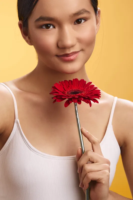 Asian woman smiles softly while holding a red flower close to her chest against a warm yellow backdrop, exuding graceful charm and quiet confidence. Her calm posture adds warmth and approachable