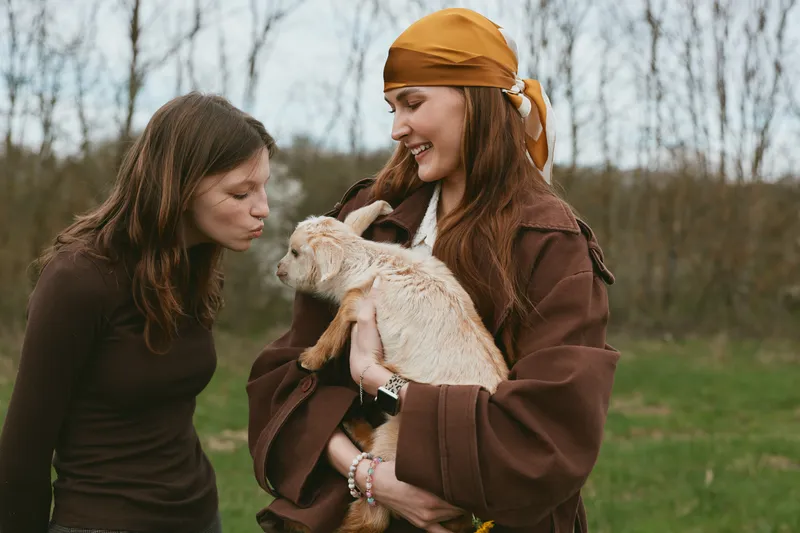 Two women smile and interact outdoors with a curious goat kid, capturing everyday friendship and lifestyle in a rural nature setting, with soft light and warm, caring moments.