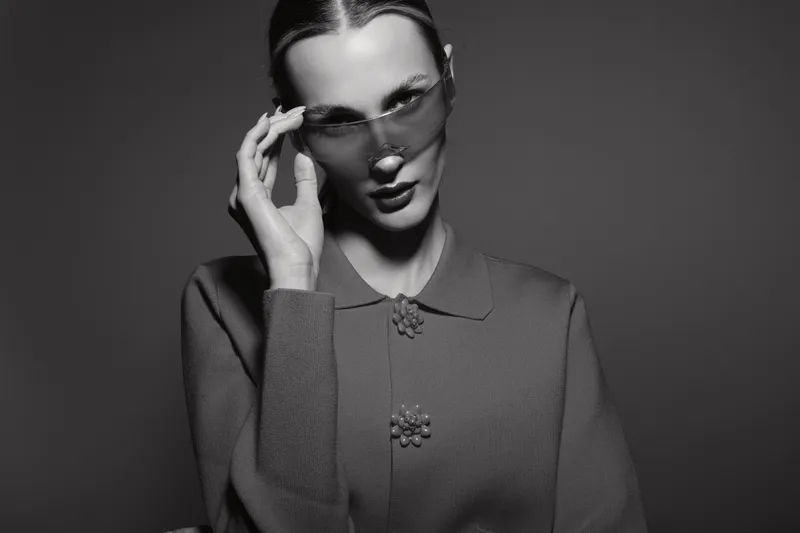 Portrait of a young white woman wearing transparent glasses and a textured buttoned sweater, posing against a dark background with a serious expression.