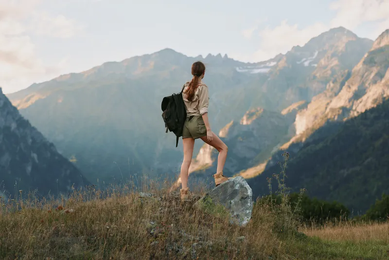 Woman hiking outdoors with backpack in a mountain landscape, capturing a sense of adventure, balance, and nature in a sunlit breeze and expansive scenery
