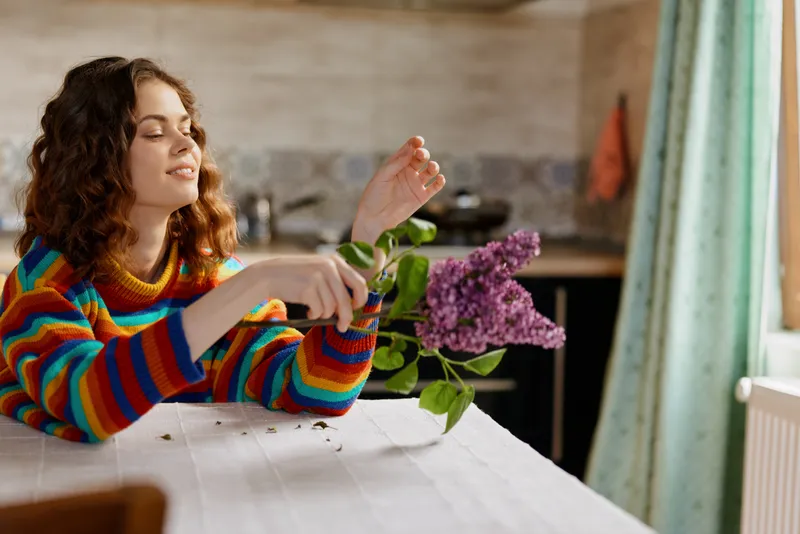Woman at a bright kitchen table arranges a bouquet of lilac flowers, exuding a casual home lifestyle with natural light and a relaxed, candid mood