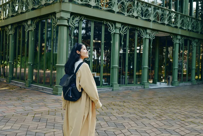 Woman with backpack in a park near ornate green architecture, outdoor stroll, coat and casual style, showcasing travel vibe, urban scenery and daylight calm.