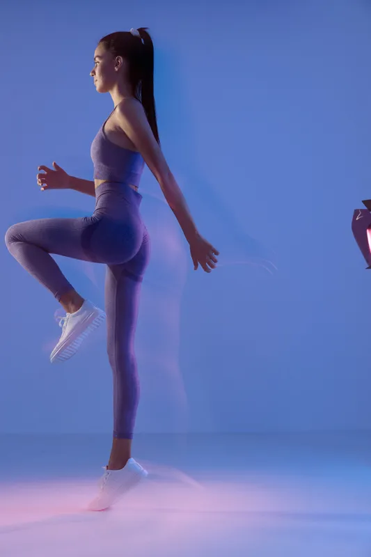 athletic woman sport fitness performing high knee exercise in purple activewear with ponytail, focused fitness training in studio with blue and pink lighting.