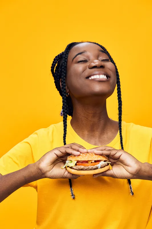 Joyful young woman enjoying a delicious burger against a vibrant yellow background, highlighting the joy of healthy eating with colorful ingredients