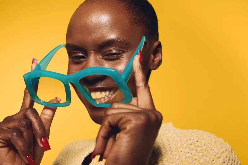 Happy dark skinned African woman with short hair holds oversized blue glasses near face smiling against yellow background. Concept of joy, fashion, beauty, and playful mood with vibrant colors.