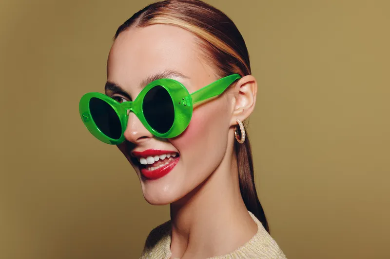 Smiling woman with green oversized sunglasses and red lipstick poses against beige background. Fashion portrait with trendy accessories and joyful expression in studio lighting.