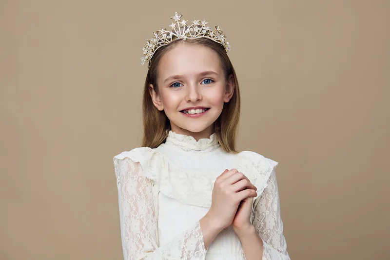 A young girl wearing a tiara and a white lace dress, smiling sweetly against a soft beige background, capturing a joyful moment of innocence and charm
