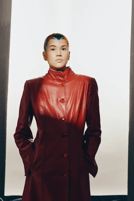 Woman with short colorful hair wearing a long red leather coat stands confidently in a bright studio with hands in pockets, showcasing fashion and modern style.