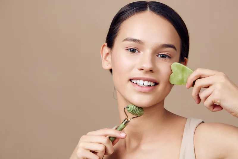 Young woman performing facial massage with jade roller and gua sha tool, promoting healthy skin and beauty routines, minimalistic background in warm tones