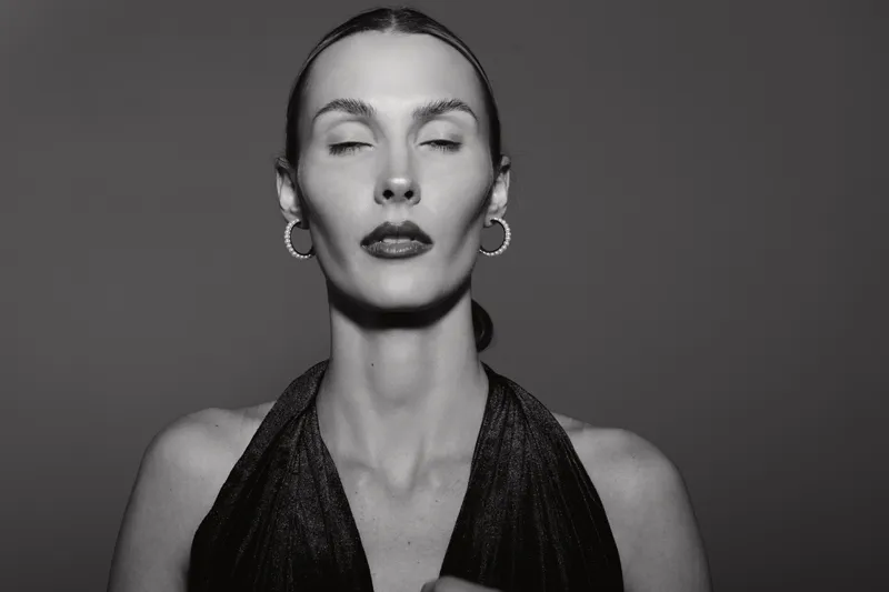 Elegant Caucasian woman with pale skin and closed eyes wears black halter dress and hoop earrings. Studio portrait in black and white with soft lighting and minimalistic background.