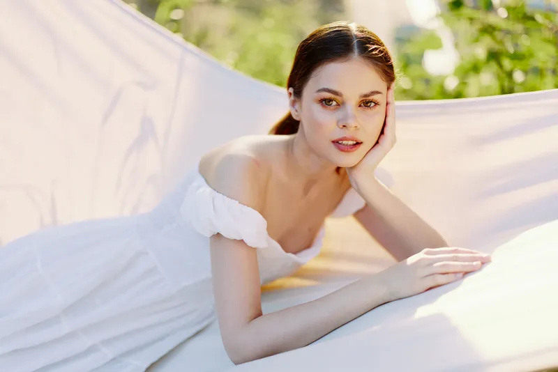 Young woman in a white dress resting on a hammock, showcasing a serene expression in a sunlit outdoor setting, embodying tranquility and relaxation