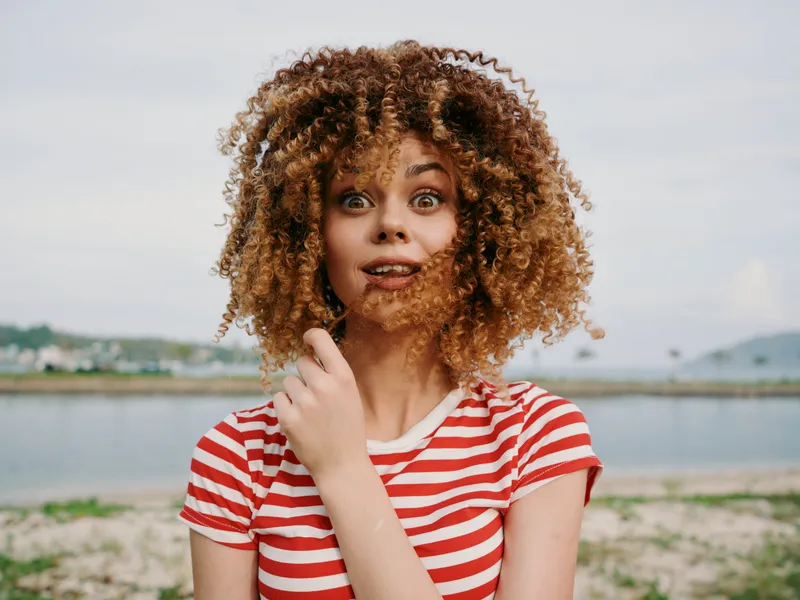 Young woman with curly hair in a red striped tee stands on a sunny beach, wide eyes and playful smile framed by wind lifting her hair.