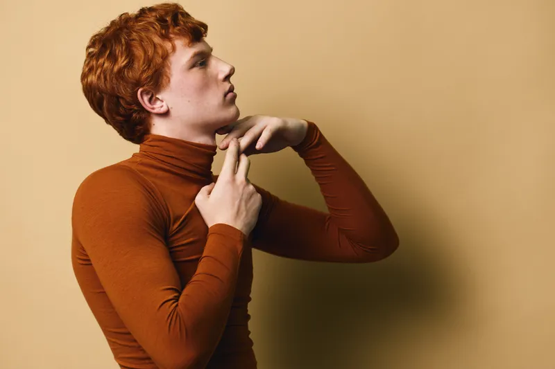 Young man with red hair wearing brown turtleneck posing on beige background, side profile view with thoughtful expression and folded hands near neck.