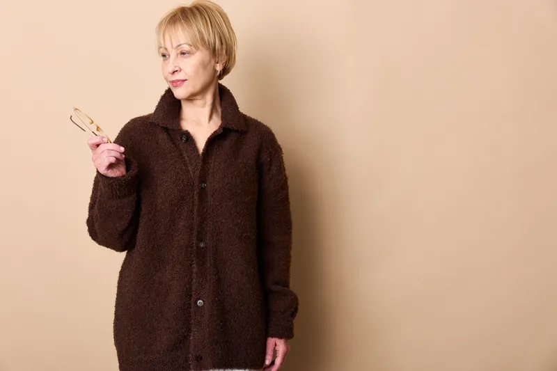 Senior woman in beige studio portrait wearing cozy jacket, thinking with hand gesture and puzzled expression, casual fashion concept for hesitation and doubt