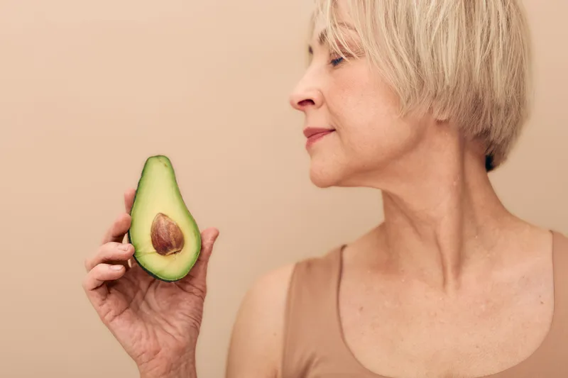 Mature woman with short blonde hair holds half of fresh avocado close to her face on beige background. Concept for healthy lifestyle, nutrition, natural beauty, and wellness at any age.