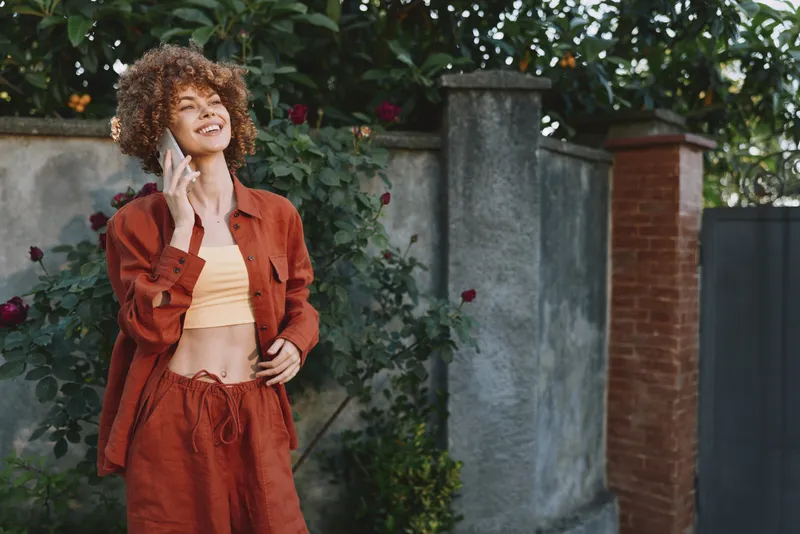 A stylish young woman with curly hair talks on a phone in a rust colored outfit in a sunny outdoor garden setting