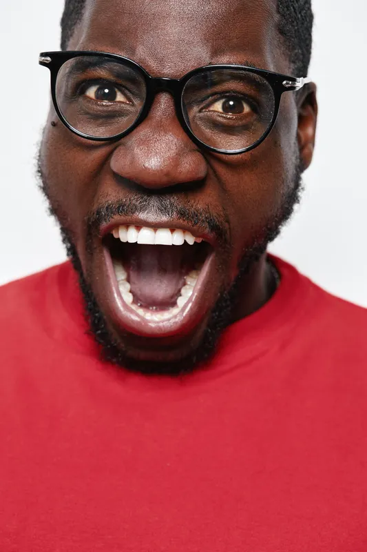 African man with dark skin and glasses wearing red shirt shouting or expressing excitement with open mouth on white background. Closeup portrait of emotional black male with facial hair.