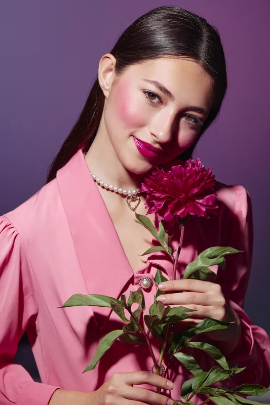 Young woman in a pink structured blazer holding a deep magenta flower. Creative beauty and fashion portrait with striking blush and glossy lip makeup on a purple background.