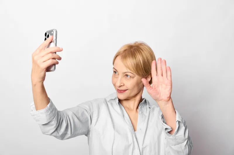 Woman holding smartphone for selfie, waving with hand gesture near ear, studio portrait on plain light background, calm listening mood, soft studio lighting