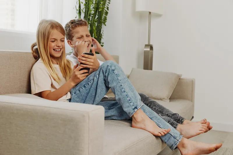 Happy siblings sitting barefoot on a couch, smiling and enjoying content on a smartphone in a bright living room. Concept of family bonding, technology, and leisure time.
