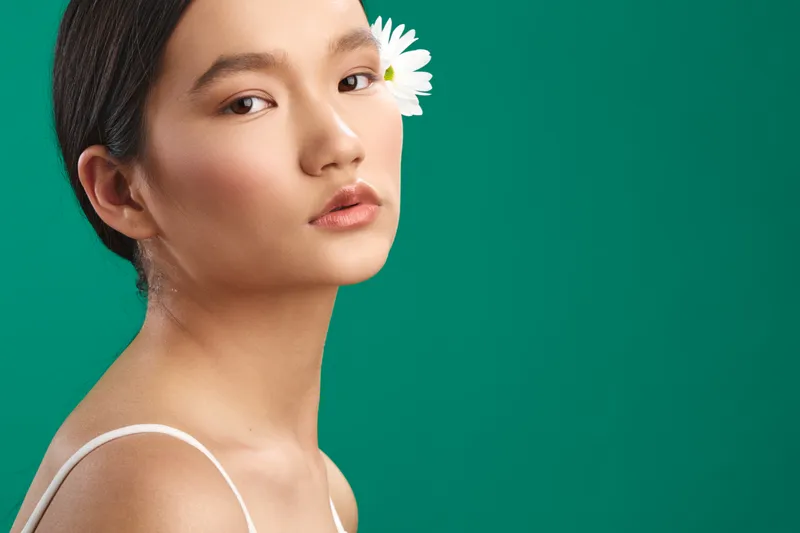 asian woman with a flower tucked behind her ear poses against a vibrant green backdrop, showcasing calm beauty and graceful features in a clean studio portrait.