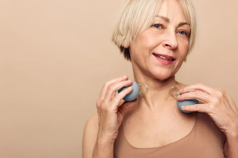 Mature woman with short blonde hair uses silicone suction cups on neck, performing facial massage in beige studio. Concept of anti aging skincare, self care, and natural beauty treatment.