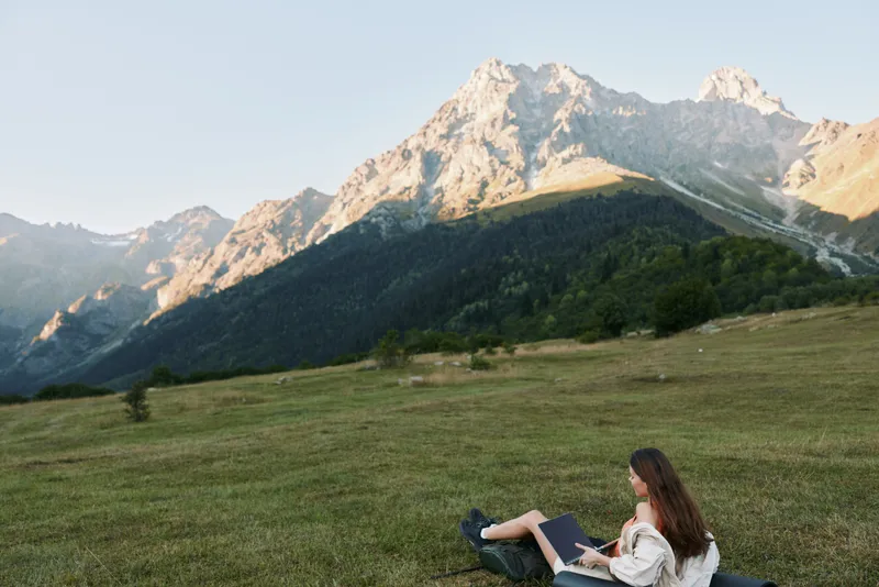 A person sits on a grassy meadow with a laptop, mountains in the distance, enjoying outdoor study and creative work in a peaceful natural setting.