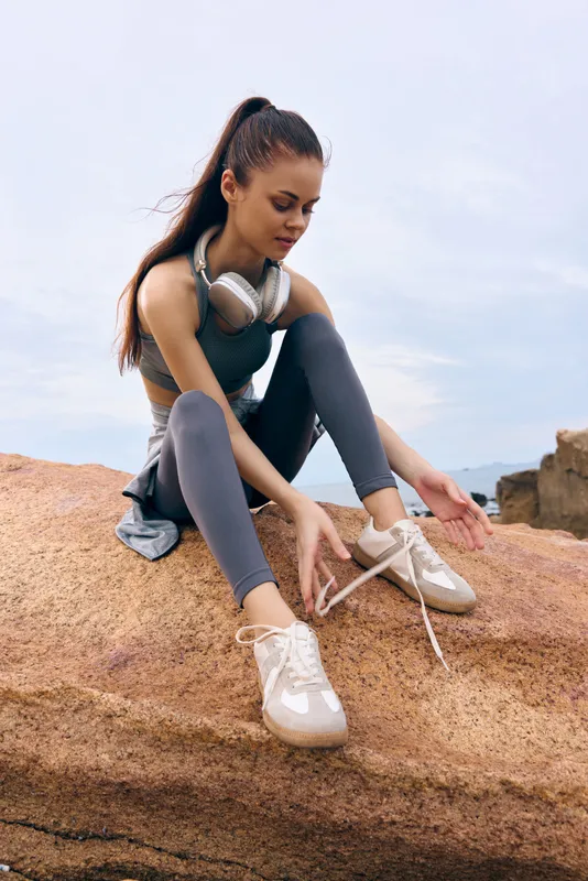 Young woman in athletic wear tying her shoelaces on a rock, showcasing an active lifestyle and fitness motivation, with natural outdoor background, bright colors, and casual style