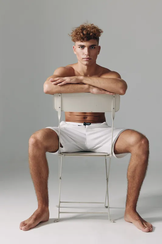 Young man with curly hair sitting barefoot on chair backwards wearing white shorts against gray background in studio. Casual pose with arms resting on chair back.