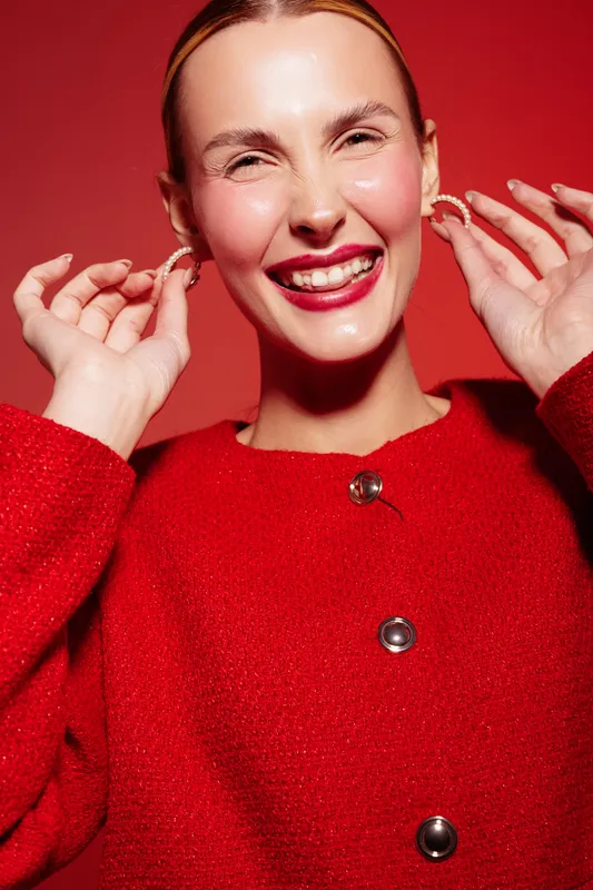 Smiling woman in red sweater holds earrings near face against red background. Close-up portrait with bright makeup and joyful expression. Concept of fashion, beauty, and accessories.
