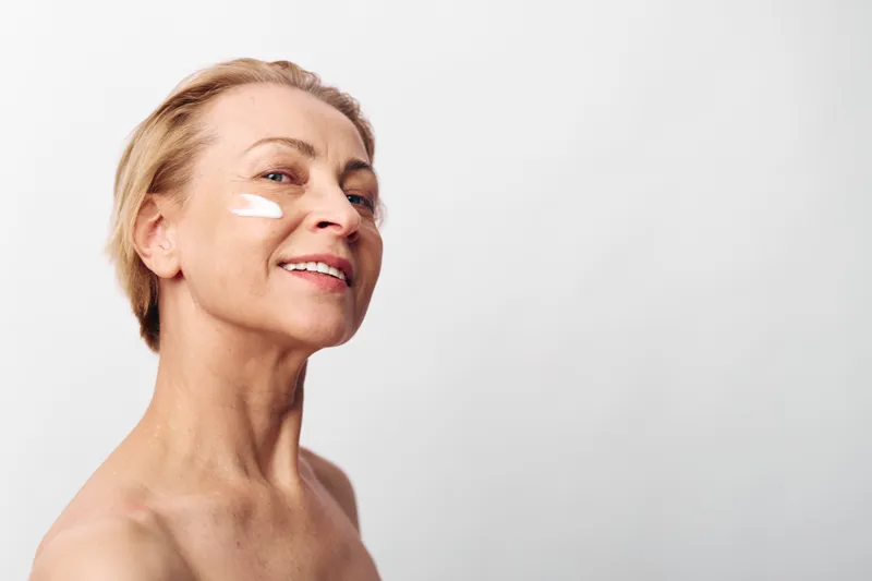 Smiling mature woman with short blonde hair applies white face cream on cheek. Clean skin, skincare concept with natural beauty and self-care isolated on white background in studio light.