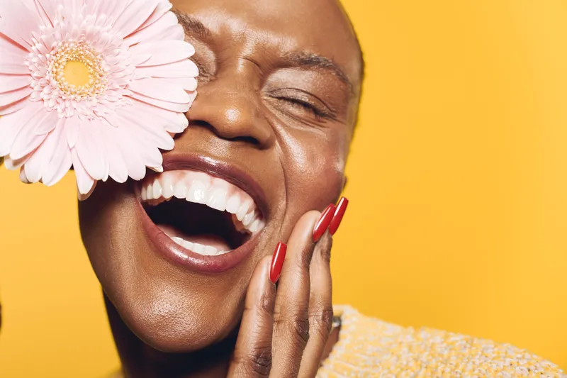 Joyful black woman with dark skin laughs with closed eyes holding pink flower near face on yellow background. Concept of happiness, beauty, natural skincare, and floral wellness.