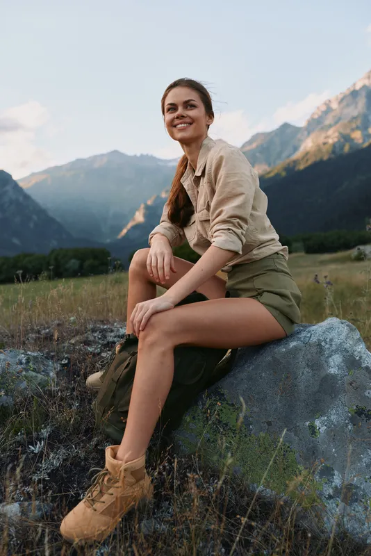 Young woman enjoys a relaxed lifestyle outdoors, sitting on a rock with mountain scenery, casual outfit, warm light, content expression, connection with nature, fresh air.