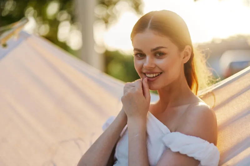 Young woman with natural beauty enjoying a sunny day, smiling softly in a hammock, dressed in a white off shoulder top, surrounded by greenery