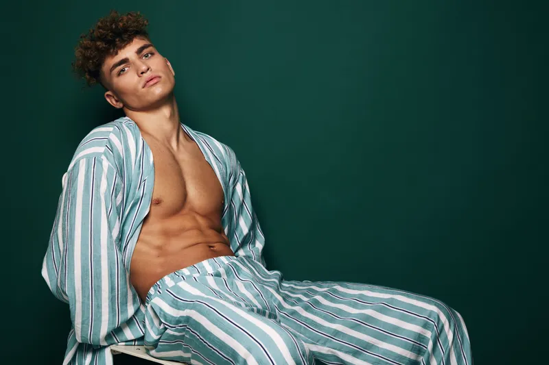 Young man with painted black nails posing in stripped loungewear with open shirt and bare chest against a dark green studio background for fashion and style concept.