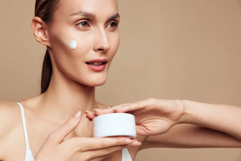 A focused woman applies moisturizer to her cheek, holding a jar while a second hand smooths cream. A clean, minimal setting highlights skincare routine and self care.